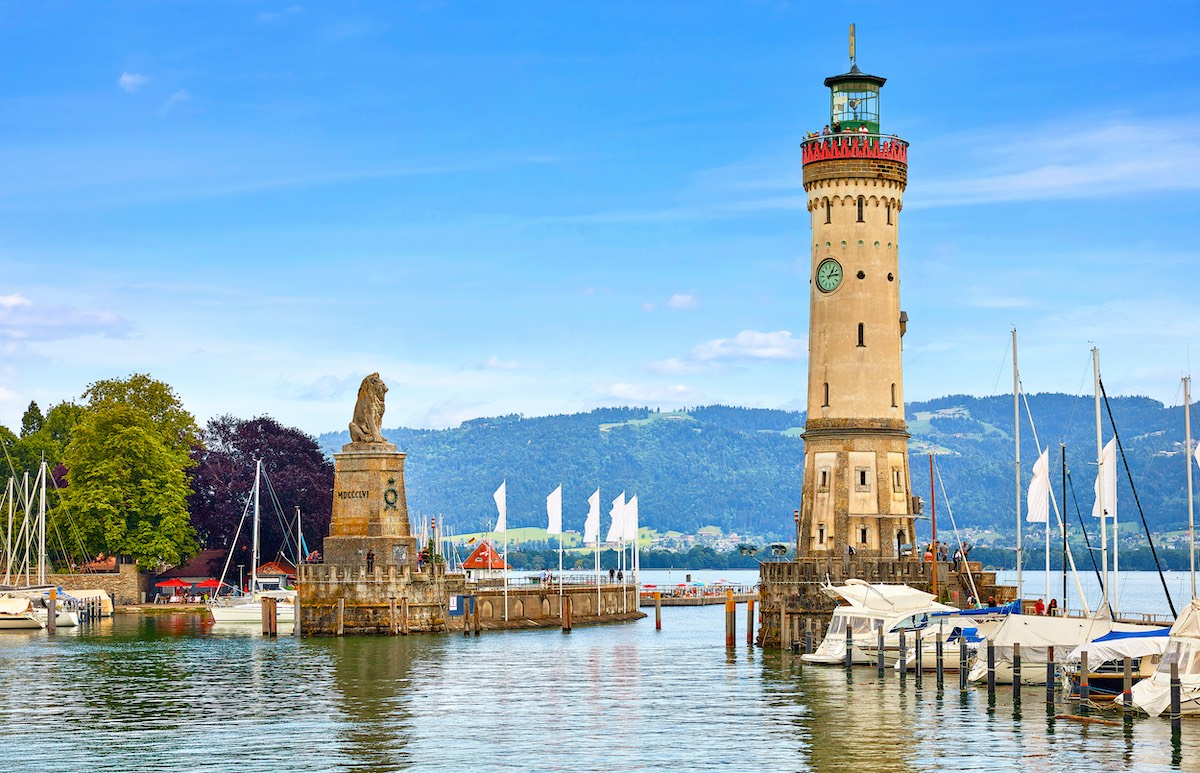 Lindau, Germany. Old lighthouse with clock in bay. Antique bavarian town at Lake Constance (Bodensee). Monument with statue of lion at entrance to port, yachts by piers. Summer landscape blue sky.